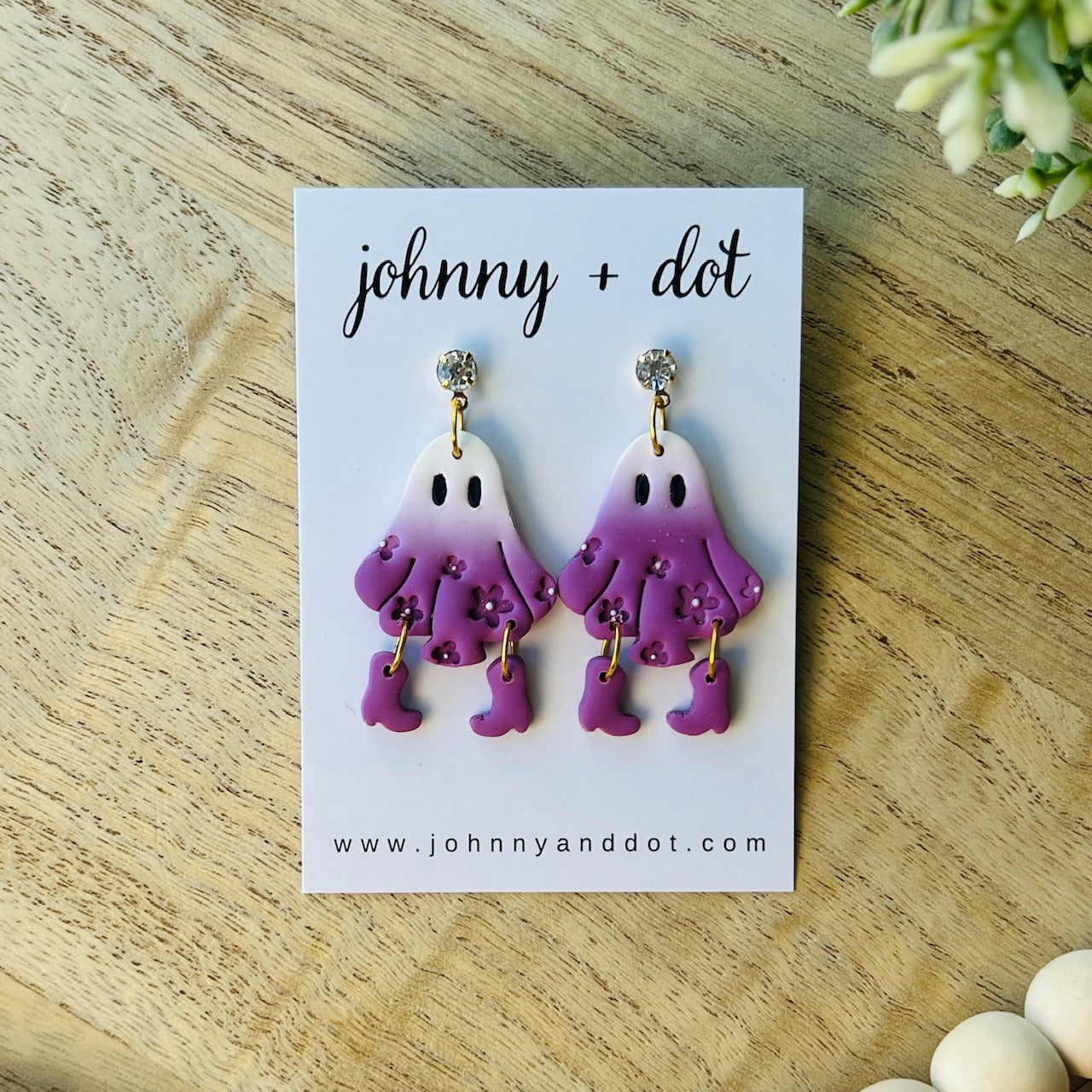 Pair of ghost-shaped earrings on a 'johnny + dot' branded card on a wooden surface.