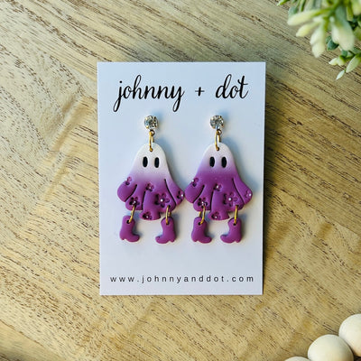 Pair of ghost-shaped earrings on a 'johnny + dot' branded card on a wooden surface.