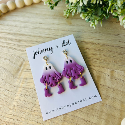 Purple ghost-shaped earrings on a 'johnny + dot' branded card with a wooden surface and flowers in the background.