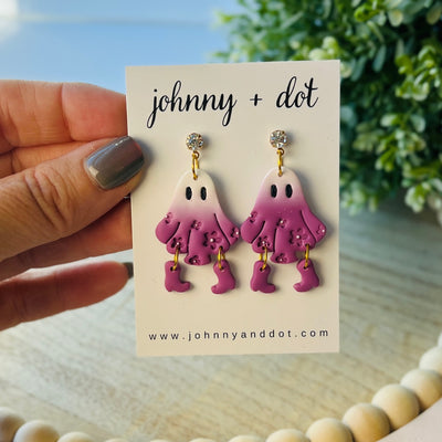 Pink ghost-shaped earrings on a 'johnny + dot' card with a blurred natural background