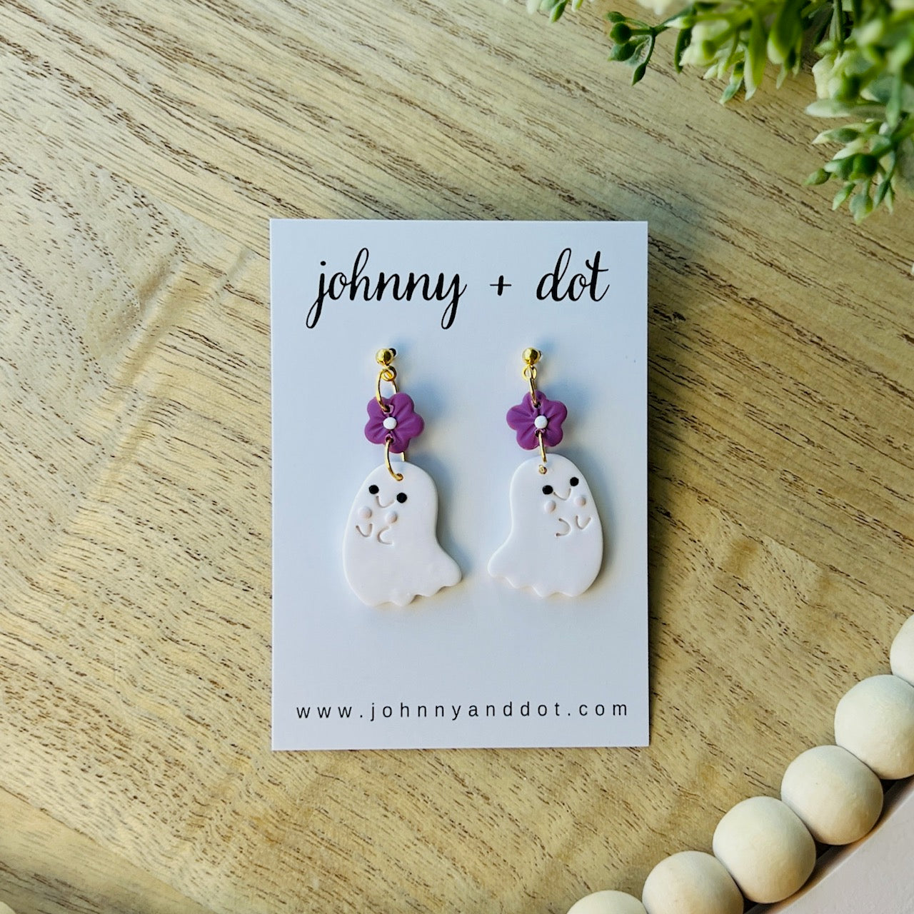 Ghost-shaped earrings with purple flowers on a 'johnny + dot' card on a wooden surface.