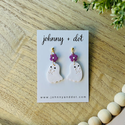 Ghost-shaped earrings with purple flowers on a 'johnny + dot' card on a wooden surface.