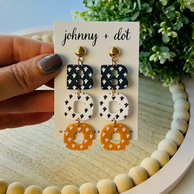 Pair of earrings on a 'johnny + dot' branded card held by a hand with a blurred background.