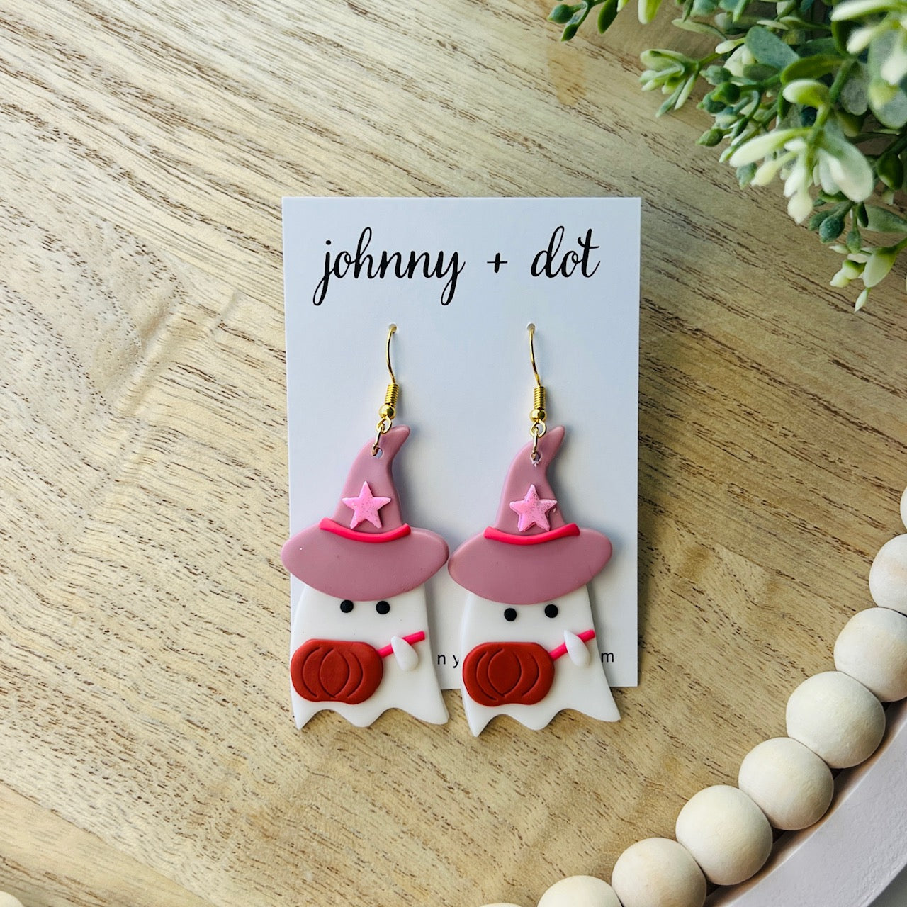 Ghost-shaped earrings with pink witch hats and pumpkin faces on a 'johnny + dot' card, placed on a wooden surface.