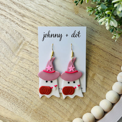 Ghost-shaped earrings with pink witch hats and pumpkin faces on a 'johnny + dot' card, placed on a wooden surface.
