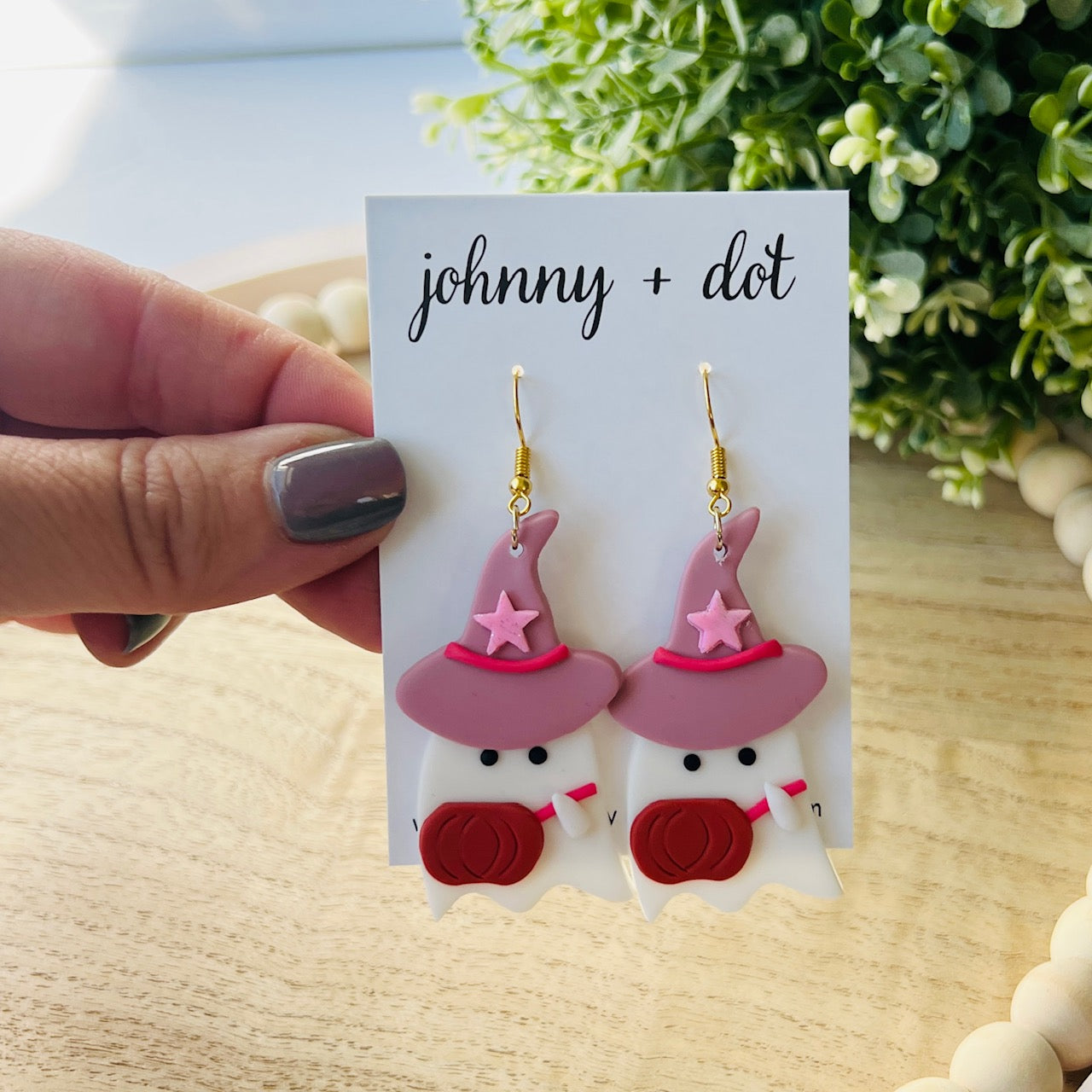 Pair of ghost earrings with pink hats and red scarves on a 'johnny + dot' card, held by a hand.