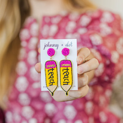 Model holding a pair of beaded earrings shaped like a pencil with the word teach written out in black beads. 