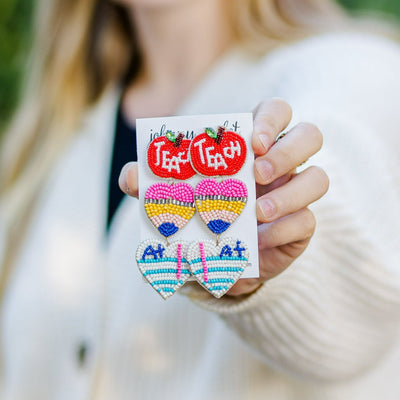 Model holding a pair of Beaded Teacher Earrings that include triple dangle of an apple, pencil, and A+ paper. 