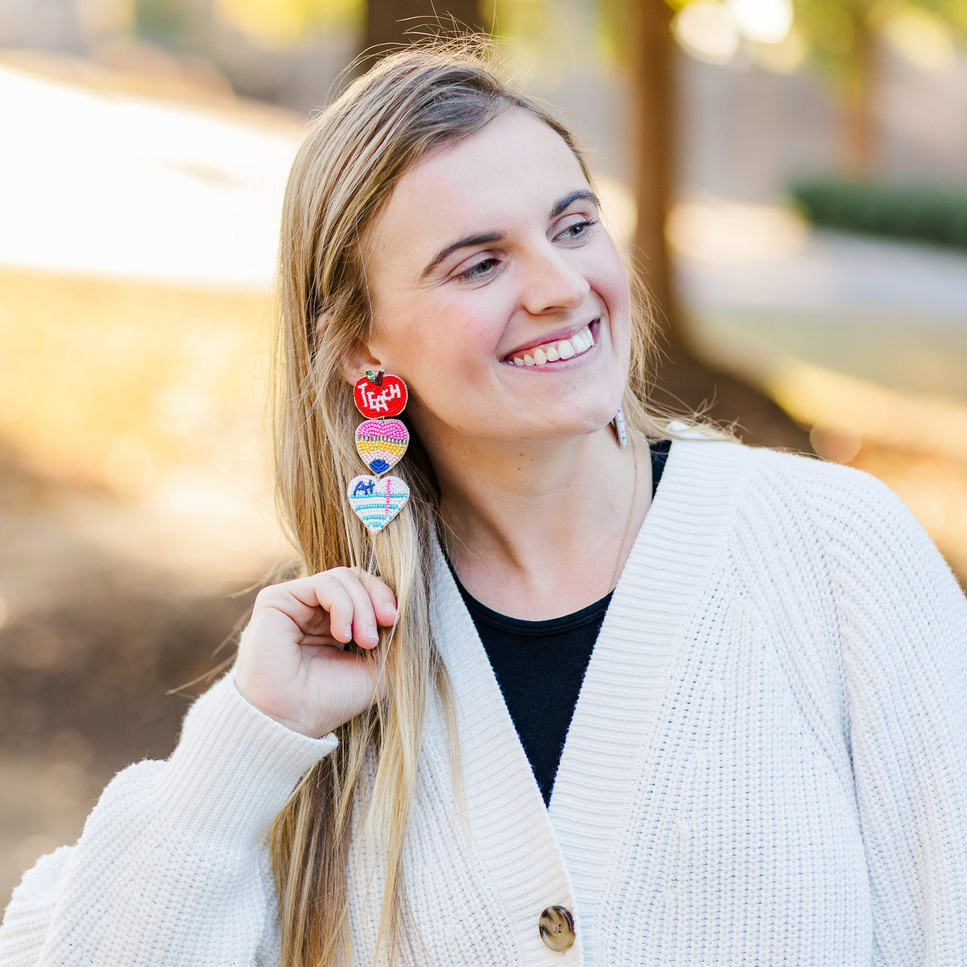 Model wearing a pair of large Beaded Teacher Earrings that include triple dangle of an apple, pencil, and A+ paper.