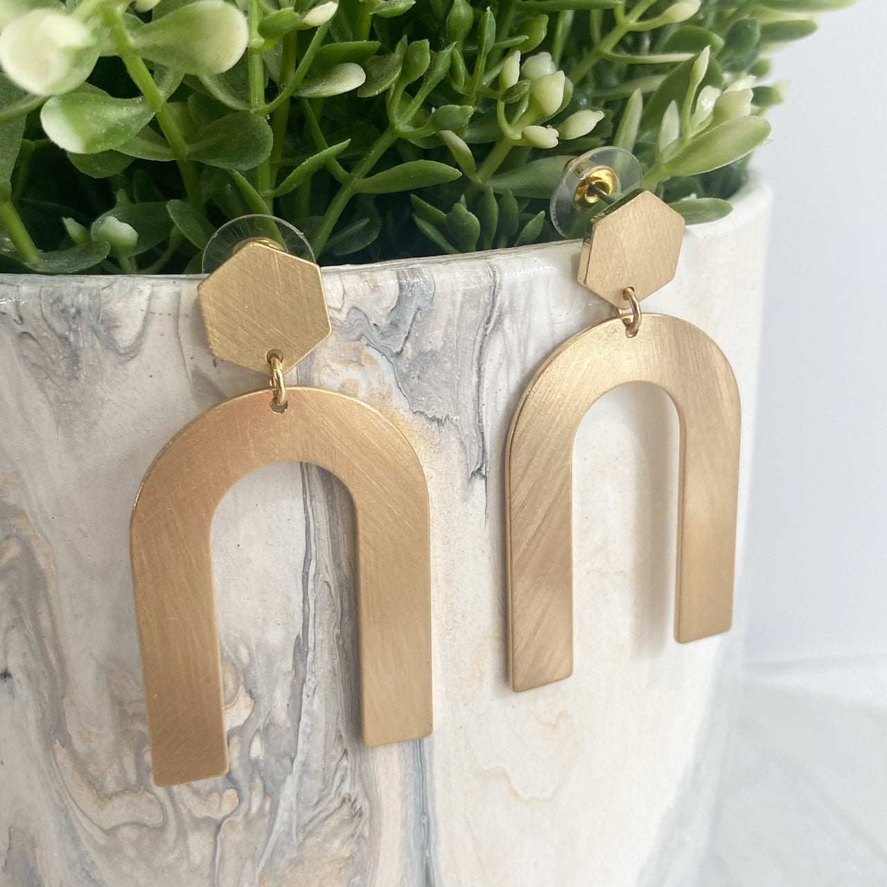 An upside u-shape metal earring with a gold metallic brushed sheen with a hexagon post-back design displayed on a faux plant.