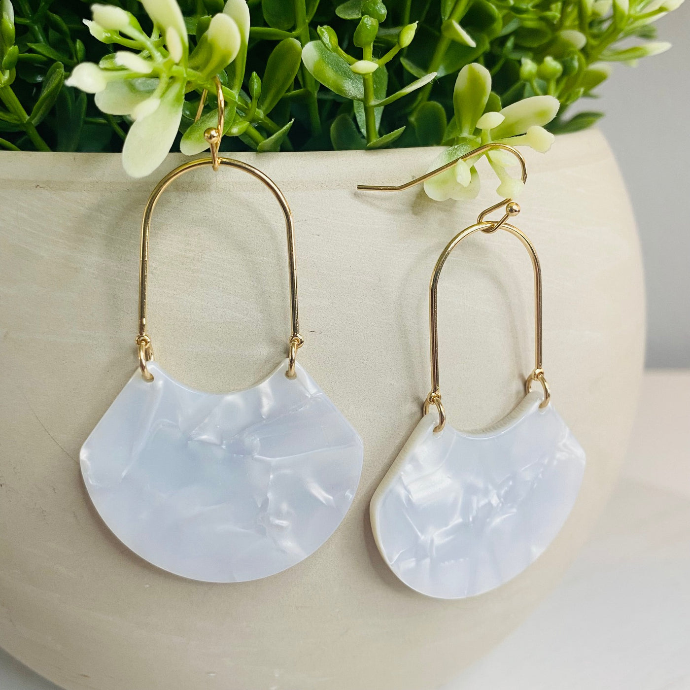 An open oval drop hoop earring with gold hardware and a white marble acetate finish displayed hung on the greenery of a faux plant.
