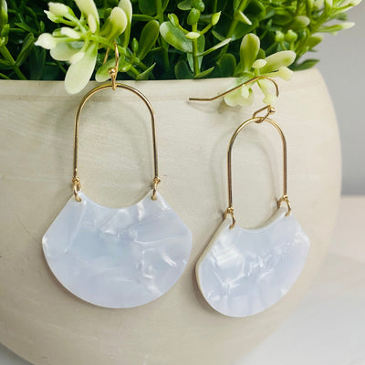 An open oval drop hoop earring with gold hardware and a white marble acetate finish displayed hung on the greenery of a faux plant.