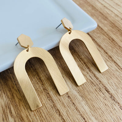 An upside u-shape metal earring with a gold metallic brushed sheen with a hexagon post-back design displayed leaning on a white plate with a wood backdrop.