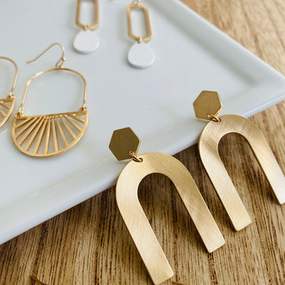 An upside u-shape metal earring with a gold metallic brushed sheen with a hexagon post-back design displayed leaning on a white plate with a few other earrings in view.